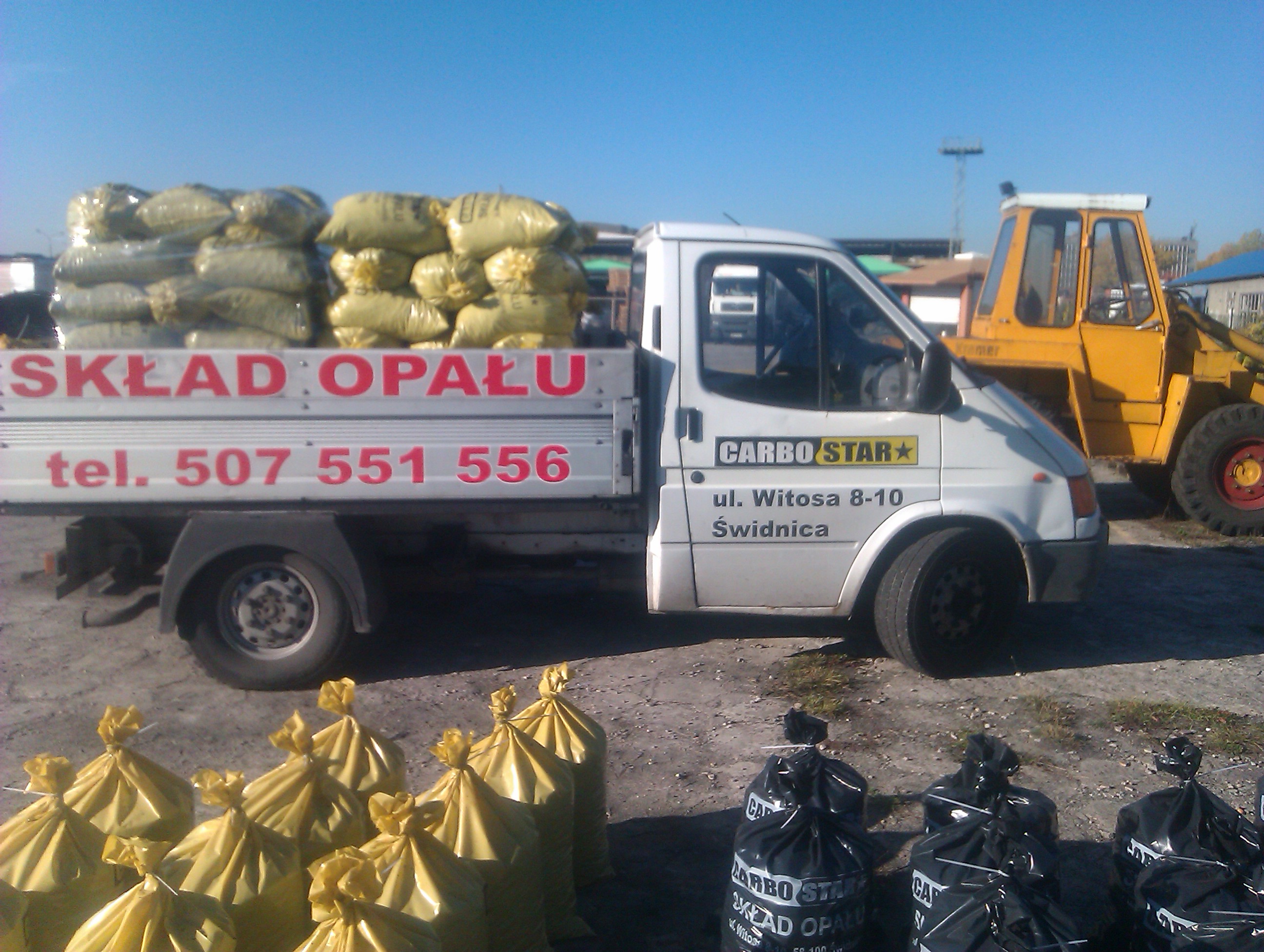 Samochód dostawczy z napisem 'Skład Opału' załadowany workami, obok stoi żółta ładowarka, a na ziemi leżą worki z opałem w kolorze żółtym i czarnym.