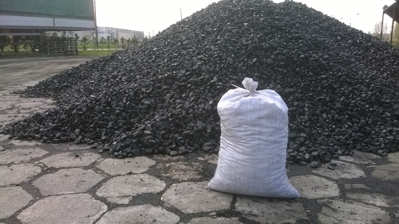 Góra czarnego węgla kamiennego na placu, obok stoi biały, wypełniony worek z węglem, widoczne płyty chodnikowe i fragment przyczepy ciężarowej w tle.