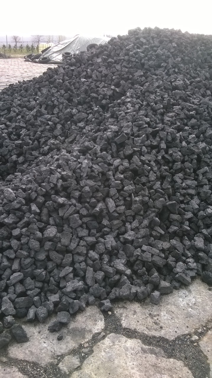 Stos czarnego koksu na betonowym podłożu z widocznymi pęknięciami, w tle przykryta plandeką sterta opon.