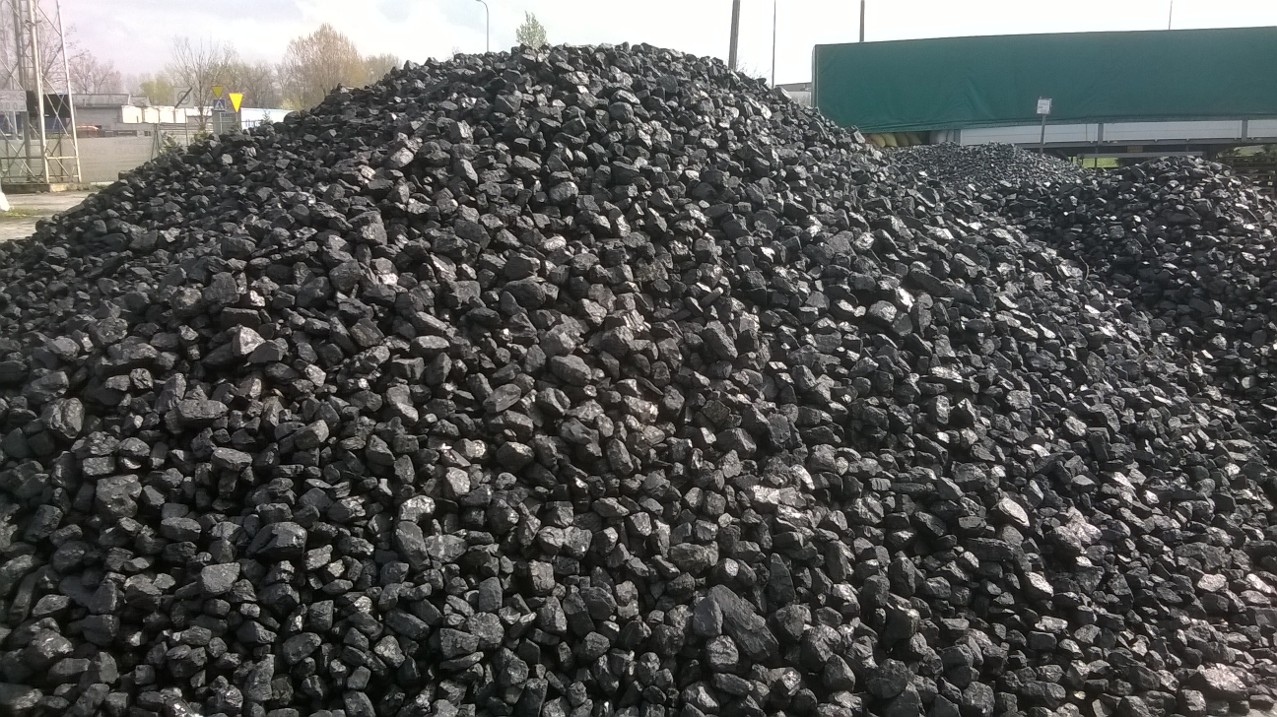 Góra czarnego węgla orzech na składzie, widoczny zielony fragment plandeki na przyczepie w tle.