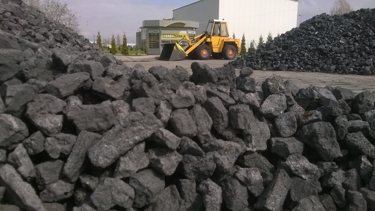 Stos koksu na placu, w tle żółta ładowarka i budynek firmy Carbo Star.