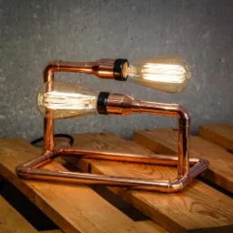 Lampa stołowa z miedzianych rur i dwóch żarówek Edisona na drewnianej palecie, z surowym betonowym tłem.