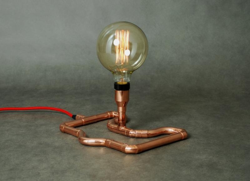 Lampa stołowa z miedzianych rurek i dekoracyjnej żarówki na szarym tle, z widocznym czerwonym kablem zasilającym.