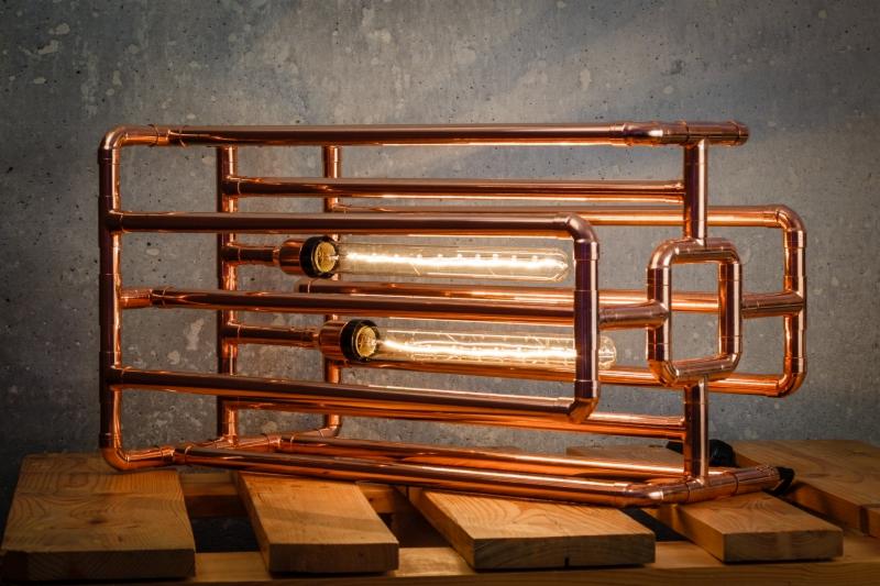 Lampa dekoracyjna wykonana z miedzianych rur i podłużnych żarówek Edisona, ustawiona na drewnianej palecie na tle szarej ściany z betonu architektonicznego.