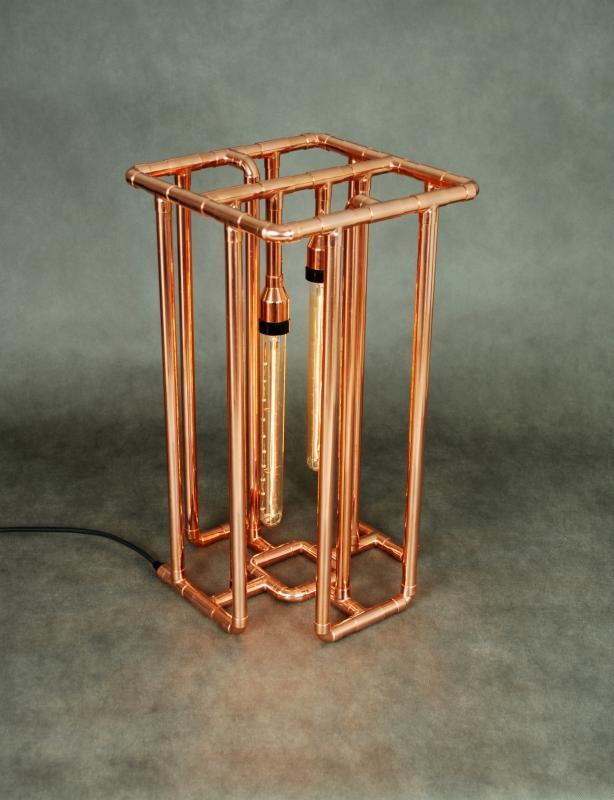 Lampa stołowa w stylu industrialnym, wykonana z miedzianych rurek, z dwiema żarówkami Edison wewnątrz klatki, na szarym tle.