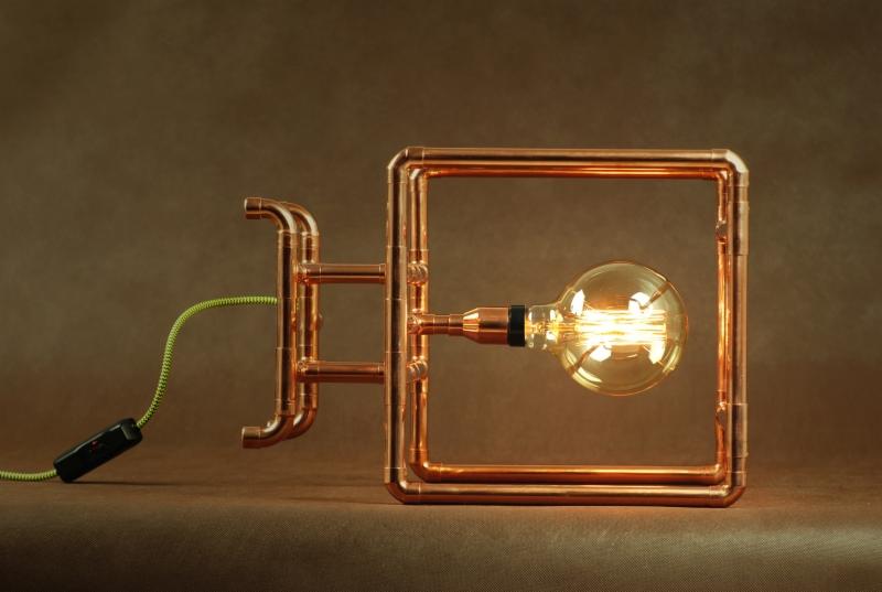 Lampa stołowa w stylu industrialnym wykonana z miedzianych rur, z żarówką Edisona i zielonym przewodem z włącznikiem, na brązowym tle.