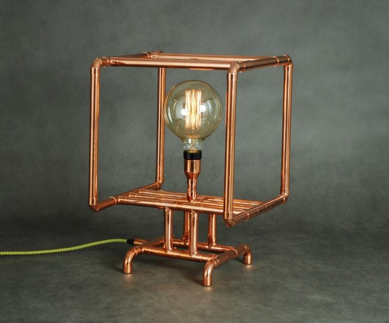Lampa stołowa w stylu industrialnym, wykonana z miedzianych rurek, z żarówką Edisona o ciepłej barwie, na szarym tle.