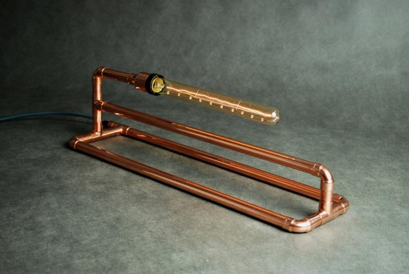 Lampa stołowa w stylu industrialnym, wykonana z miedzianych rur, z podłużną żarówką filamentową, na szarym tle.
