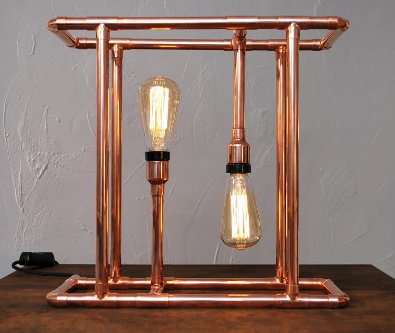 Lampa stołowa w stylu industrialnym, wykonana z miedzianych rur i dwóch żarówek Edisona, umieszczona na drewnianym blacie, z widocznym kablem zasilającym po lewej stronie.