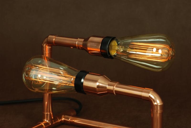 Lampa stołowa z dwoma żarówkami Edisona, wykonana z miedzianych rur i z czarnymi oprawkami, na ciemnobrązowym tle.