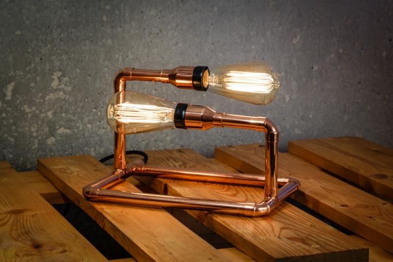 Lampa stołowa z miedzianych rur i dwóch żarówek Edisona na drewnianej palecie, z surowym betonowym tłem.