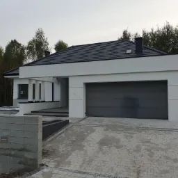 Nowoczesny dom jednorodzinny z płaskim dachem krytym ciemną dachówką, szarą bramą garażową i podjazdem wyłożonym kostką brukową w odcieniach szarości, otoczony zielenią drzew.