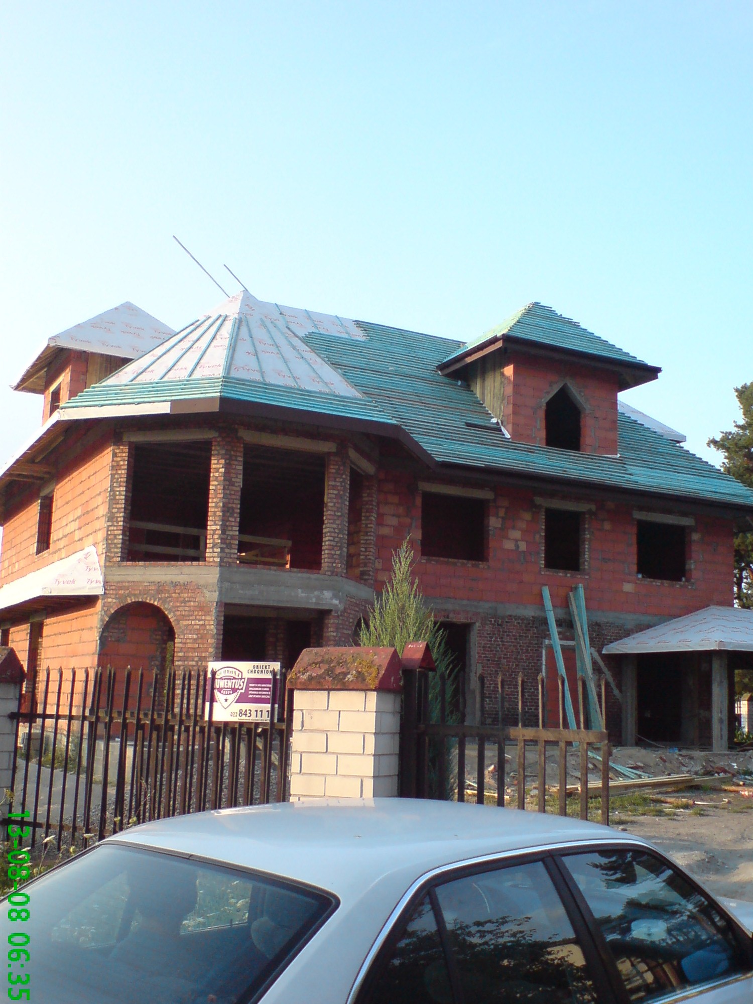 Niedokończony budynek z surowej cegły z częściowo położonym, zielonym dachem z dachówki ceramicznej, widok zza ogrodzenia i samochodu.