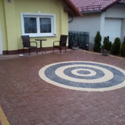 Czerwona kostka brukowa ułożona w okrągły wzór przed domem z żółtą elewacją, widoczne krzesła i stolik.