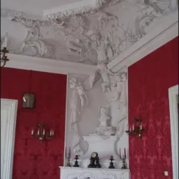 Pokój z dekoracyjnymi, czerwonymi ścianami w stylu retro, ozdobnym sufitem z białymi sztukateriami, kominkiem z rzeźbami i popiersiem oraz żyrandolami.