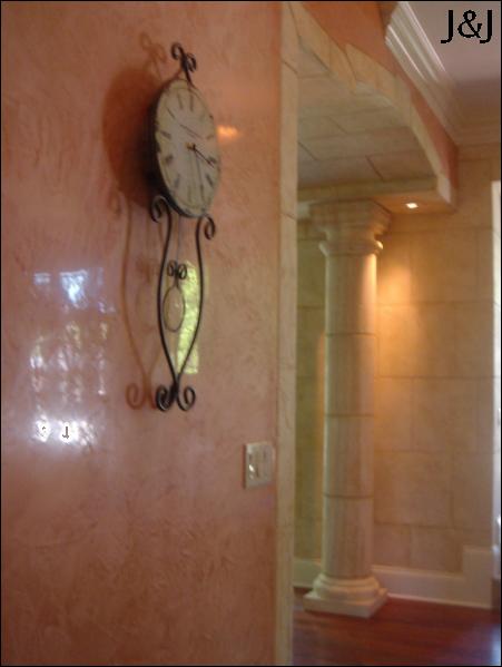 Dekoracyjna ściana w odcieniach różu z metalowym zegarem wiszącym, widoczna kolumna w przejściu do kolejnego pomieszczenia, efektowna technika malarska.