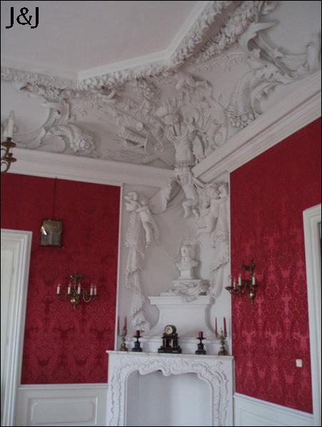 Pokój z dekoracyjnymi, czerwonymi ścianami w stylu retro, ozdobnym sufitem z białymi sztukateriami, kominkiem z rzeźbami i popiersiem oraz żyrandolami.