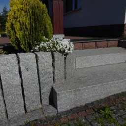 Schody zewnętrzne z szarego kamienia, otoczone zielenią i białymi kwiatami, prowadzące do domu.