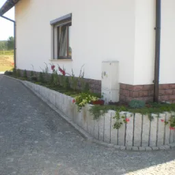 Obrzeże z pionowo ułożonych, szarych elementów kamiennych oddziela rabatę kwiatową od nawierzchni z drobnej kostki, w tle biały budynek z ceglanym cokołem.