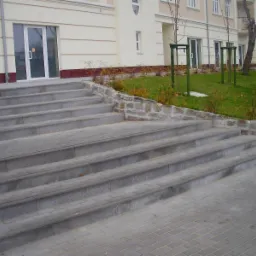 Szerokie, betonowe schody prowadzące do budynku z beżową elewacją, obok schodów niski murek z kamienia polnego i trawnik z młodymi drzewkami w zielonych osłonach.