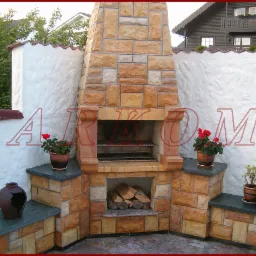 Zewnętrzny kominek z piaskowca z rusztem i zapasem drewna, otoczony białym murem i roślinami w doniczkach.