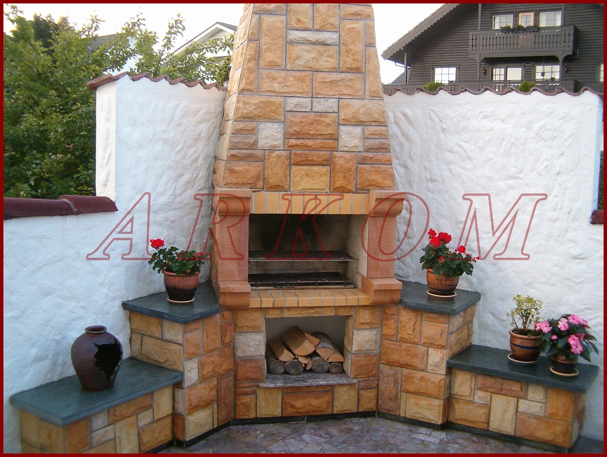 Zewnętrzny kominek z piaskowca z rusztem i zapasem drewna, otoczony białym murem i roślinami w doniczkach.