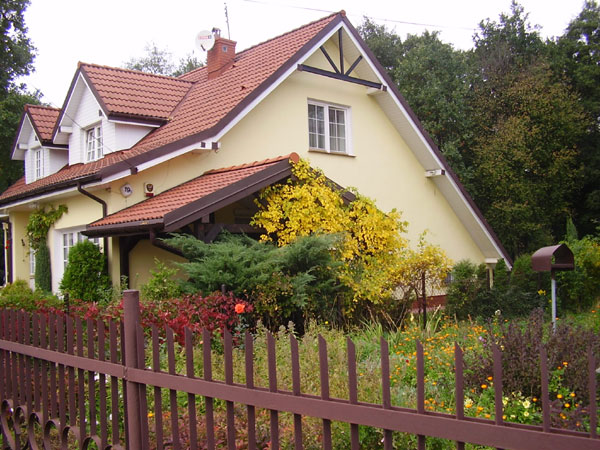 Dom jednorodzinny z czerwonym dachem z dachówki, widoczny zza ogrodzenia. Żółta elewacja, białe okna i podbitka dachowa. W tle drzewa i krzewy.
