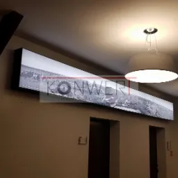 Długi, podświetlany panel z czarno-białym zdjęciem miasta, zamontowany na ścianie nad drzwiami, oświetlony przez dwie nowoczesne lampy wiszące z szarymi abażurami.