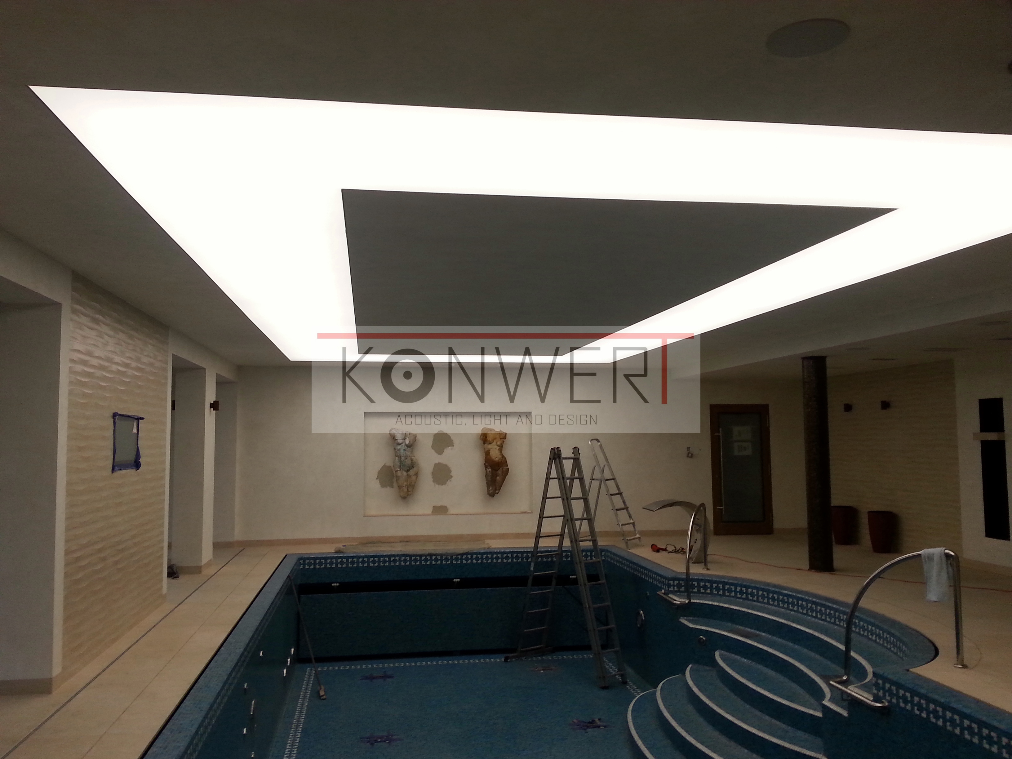 Nowoczesne oświetlenie sufitowe LED w kształcie ramy nad basenem w pomieszczeniu, dwie drabiny i rzeźby na ścianie w tle.