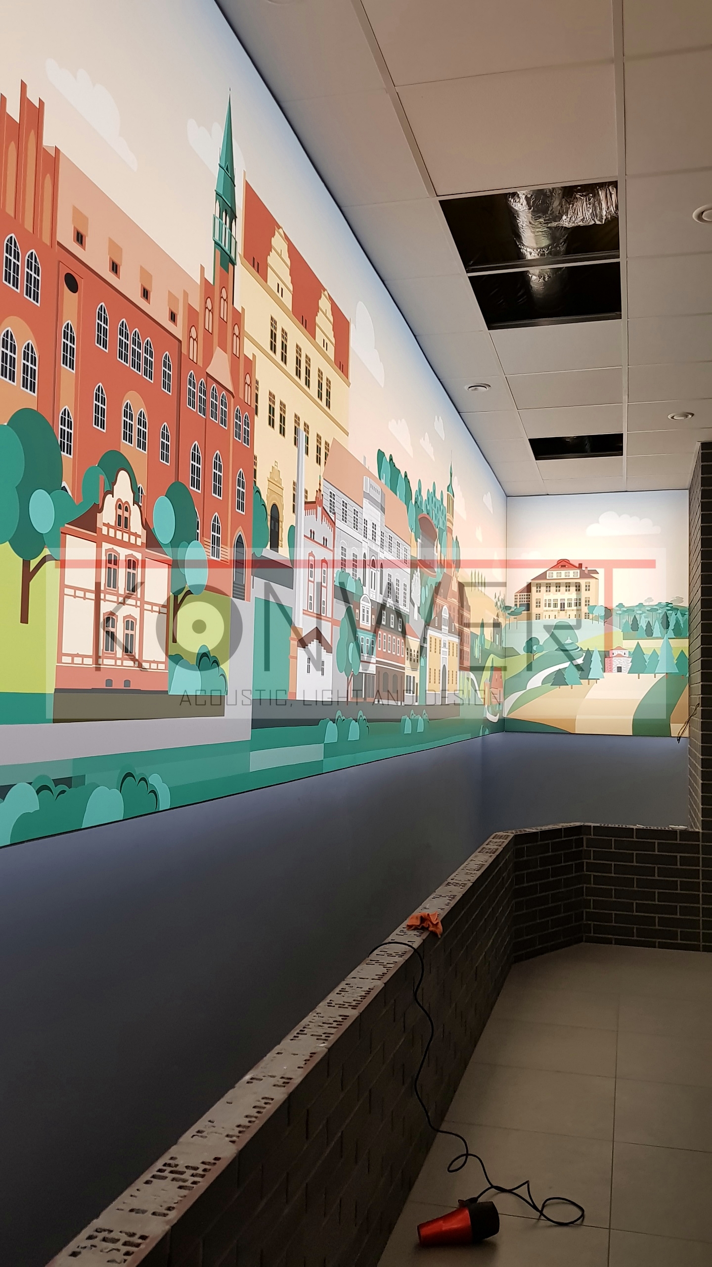 Wnętrze biura z fototapetą przedstawiającą kolorowe budynki miejskie, widoczne fragmenty sufitu podwieszanego z otworami inspekcyjnymi oraz elementy wykończenia z ciemnej cegły.