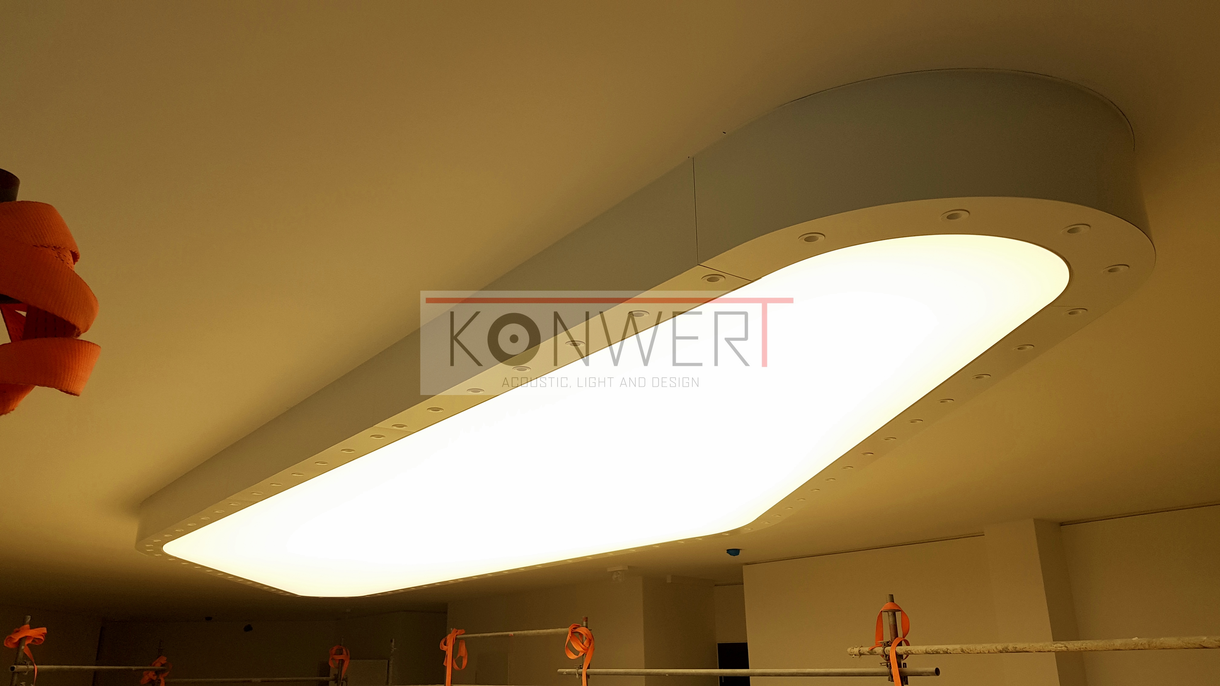 Podłużna, biała lampa sufitowa z wbudowanymi punktami świetlnymi, widoczna z dołu, z logo firmy nałożonym na zdjęcie. Fragment rusztowania z pomarańczowymi pasami w tle.