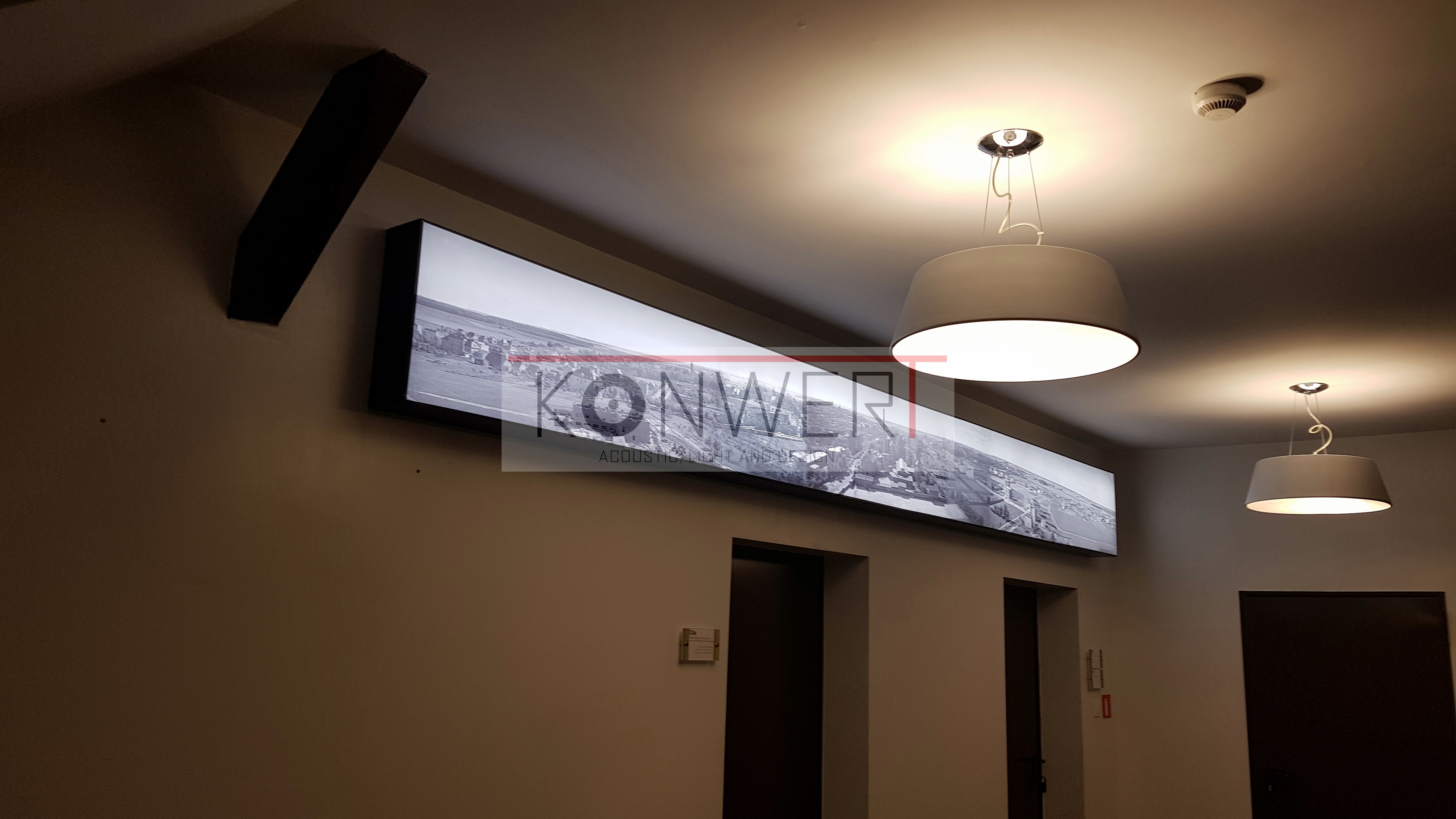 Długi, podświetlany panel z czarno-białym zdjęciem miasta, zamontowany na ścianie nad drzwiami, oświetlony przez dwie nowoczesne lampy wiszące z szarymi abażurami.