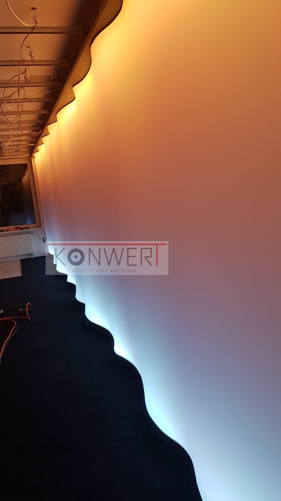 Falista listwa oświetleniowa LED, emitująca ciepłe i chłodne światło, zamontowana pod sufitem podwieszanym w nowoczesnym biurze, widoczne kable w przestrzeni nad sufitem.