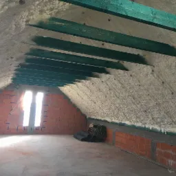 Poddasze z widoczną izolacją natryskową, zielone belki stropowe, ceglana ściana z oknem zabezpieczonym folią, zwinięte materiały izolacyjne w rogu.