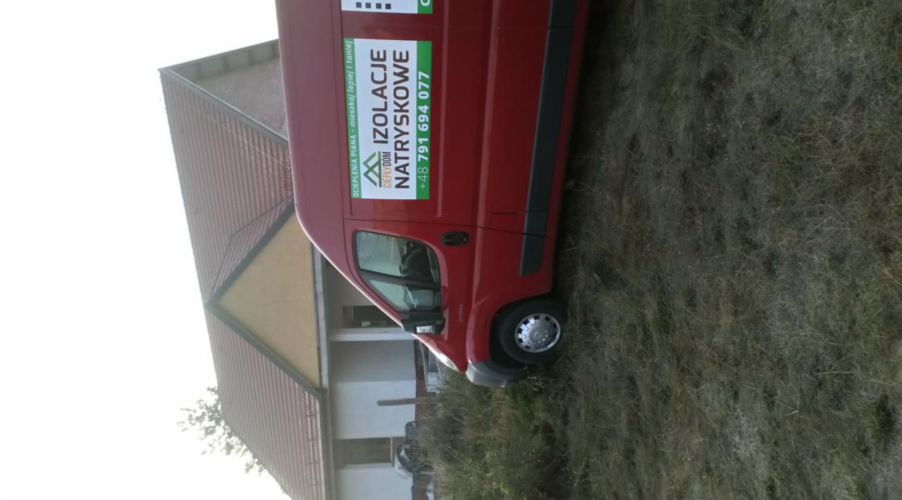 Czerwony samochód dostawczy z reklamą ocieplania pianką natryskową, zaparkowany na trawie przed domem z brązowym dachem.