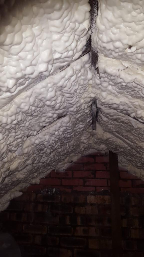 Strych z widoczną konstrukcją i zaaplikowaną warstwą izolacyjną o strukturze piany, częściowo zakrywającej ceglany mur.