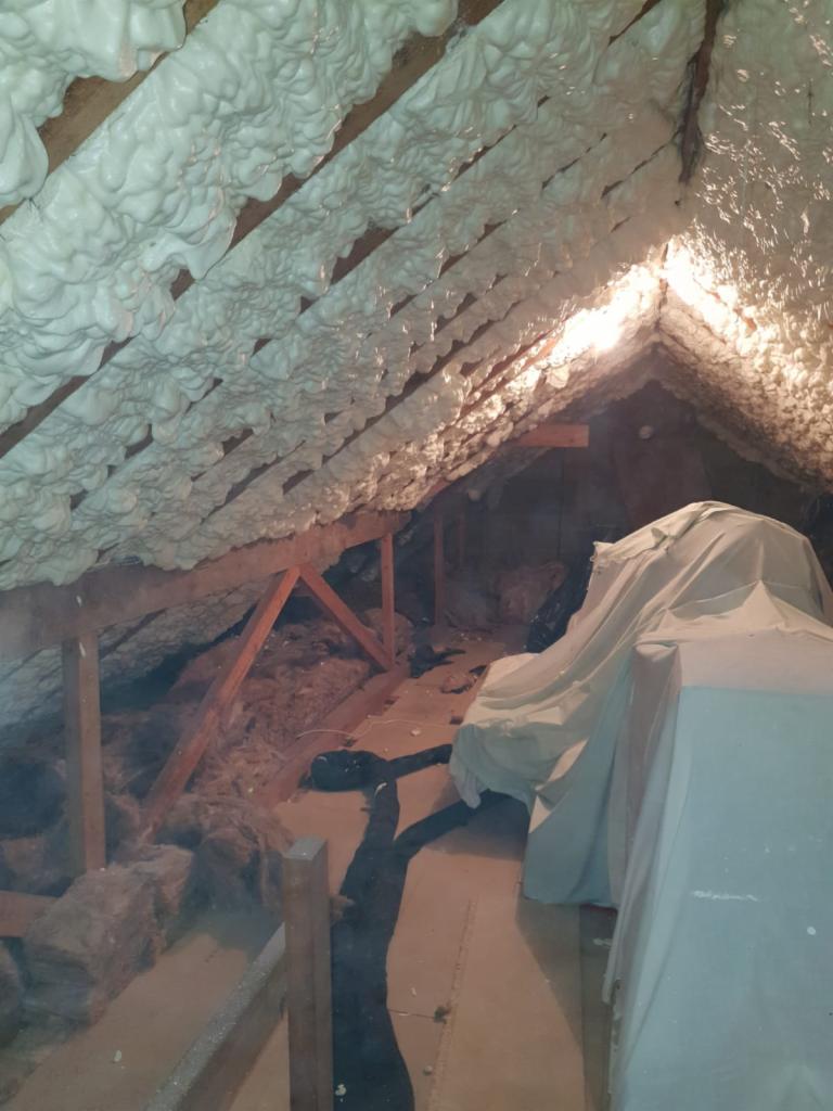 Poddasze z drewnianą konstrukcją, pokryte białą, spienioną izolacją, z widocznymi fragmentami starej wełny mineralnej i zabezpieczonymi elementami pod folią.