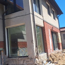 Pil-dom,Paweł Subotkiewicz - Budynek w trakcie prac elewacyjnych z widocznym fragmentem ściany z cegły, nowymi oknami i przygotowanym terenem. Prace w toku, widoczne materiały budowlane.