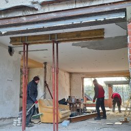 Pil-dom,Paweł Subotkiewicz - Rozbudowa domu z czerwonej cegły: robotnicy przy pracy, widoczne podpory stropu i częściowo usunięta ściana. W tle wnętrze z meblami i oknem.