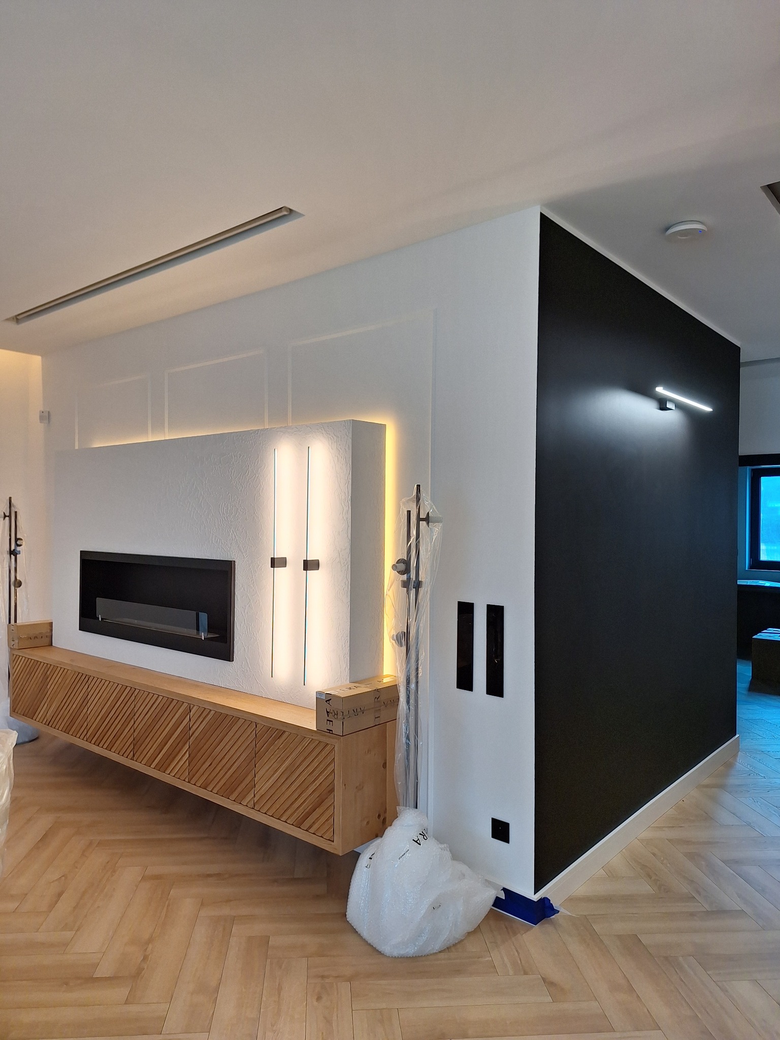 Nowoczesny salon z drewnianą podłogą w jodełkę, białą ścianą z wbudowanym kominkiem i podświetleniem LED oraz czarną ścianą akcentującą, częściowo zakryty folią ochronną.