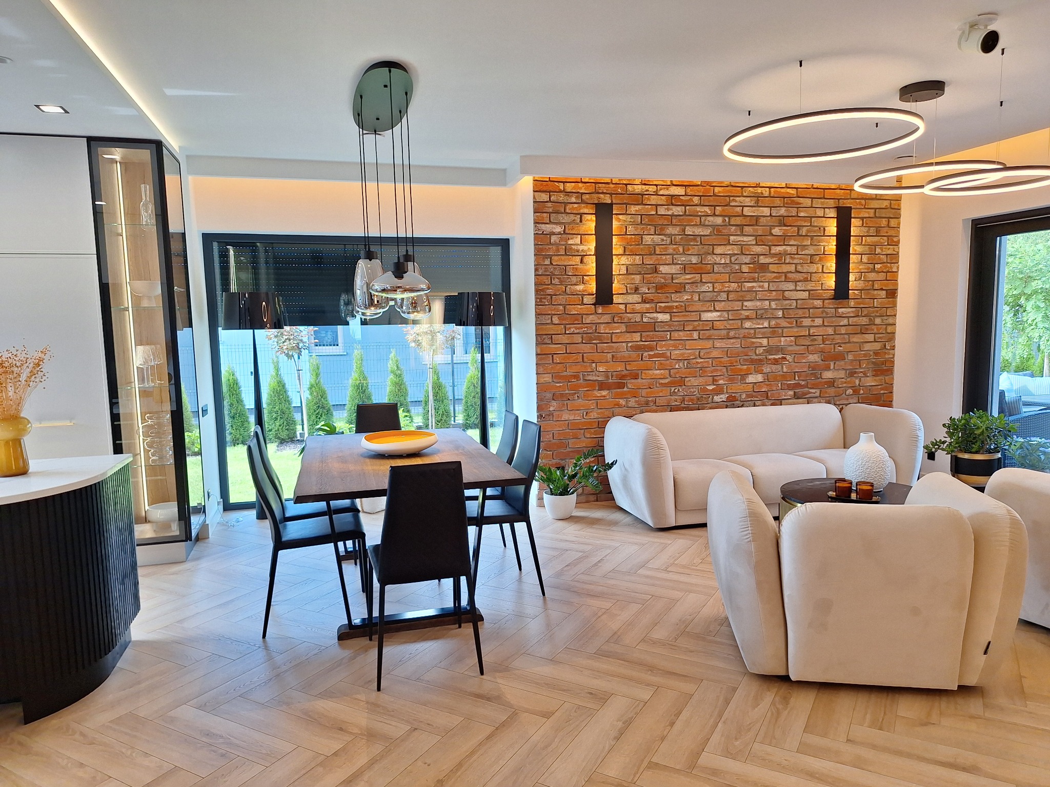 Jasny salon z ceglaną ścianą akcentującą, parkietem ułożonym w jodełkę, nowoczesnym oświetleniem i minimalistycznymi meblami w odcieniach beżu i czerni.