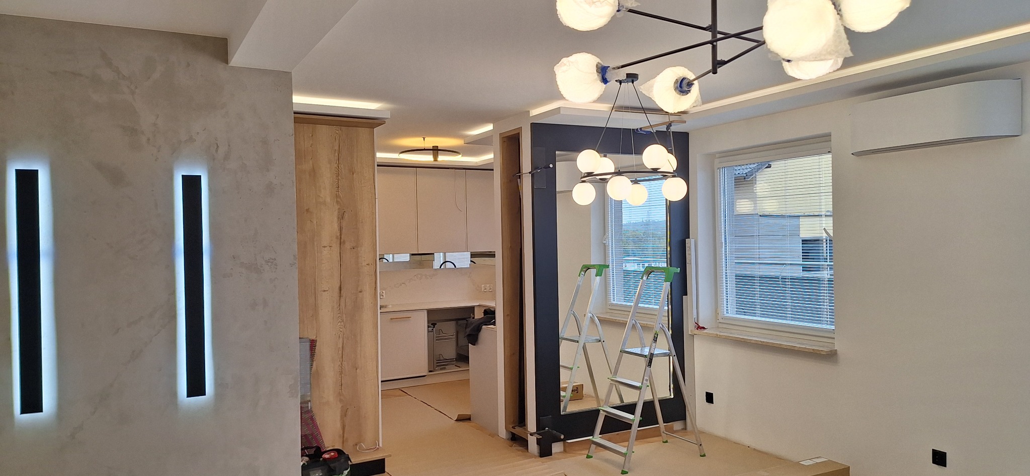 Wnętrze w trakcie wykańczania: szpachlowana ściana z podświetlanymi listwami, kuchnia w tle, drabina oparta o lustro, lampy wiszące z kulistymi kloszami w folii ochronnej.