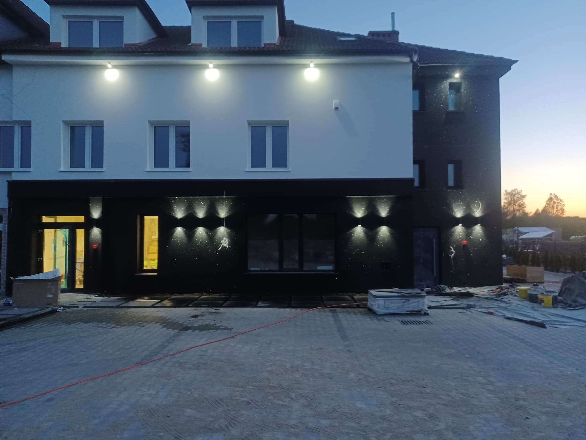 Dwukondygnacyjny budynek z jasną elewacją na górze i ciemną na dole, oświetlony zewnętrznymi lampami, z kostką brukową na podjeździe i elementami budowlanymi wokół.