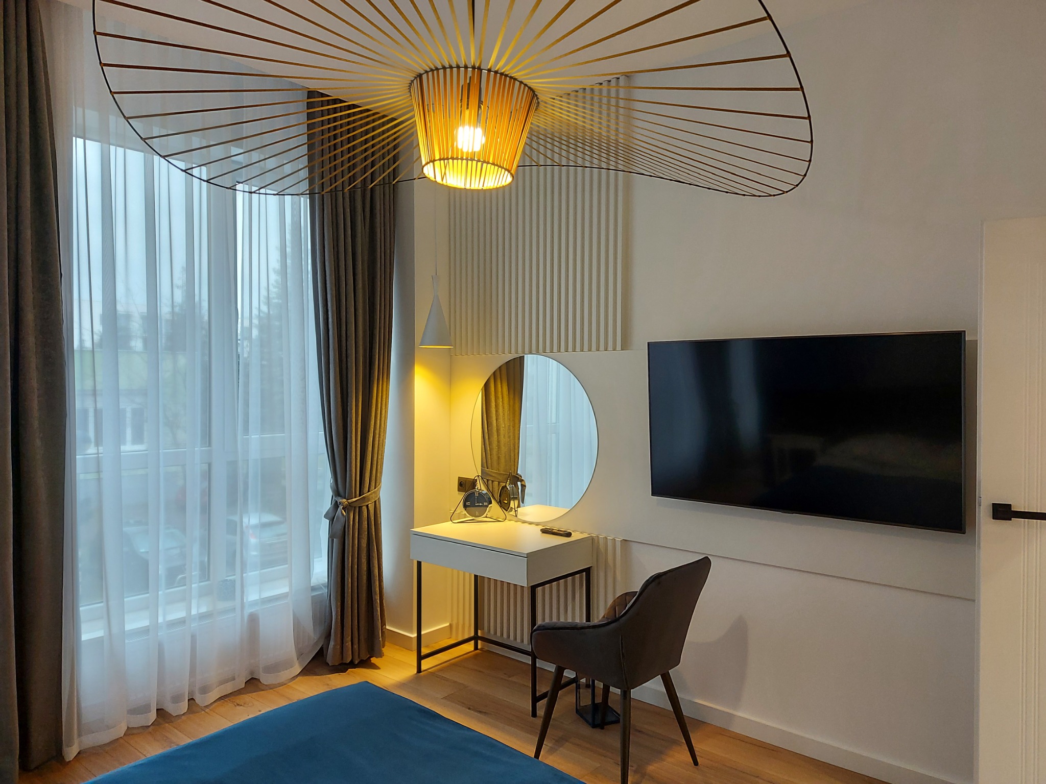 Nowoczesna sypialnia z toaletką, okrągłym lustrem, telewizorem na ścianie i designerską lampą sufitową w kształcie wachlarza.