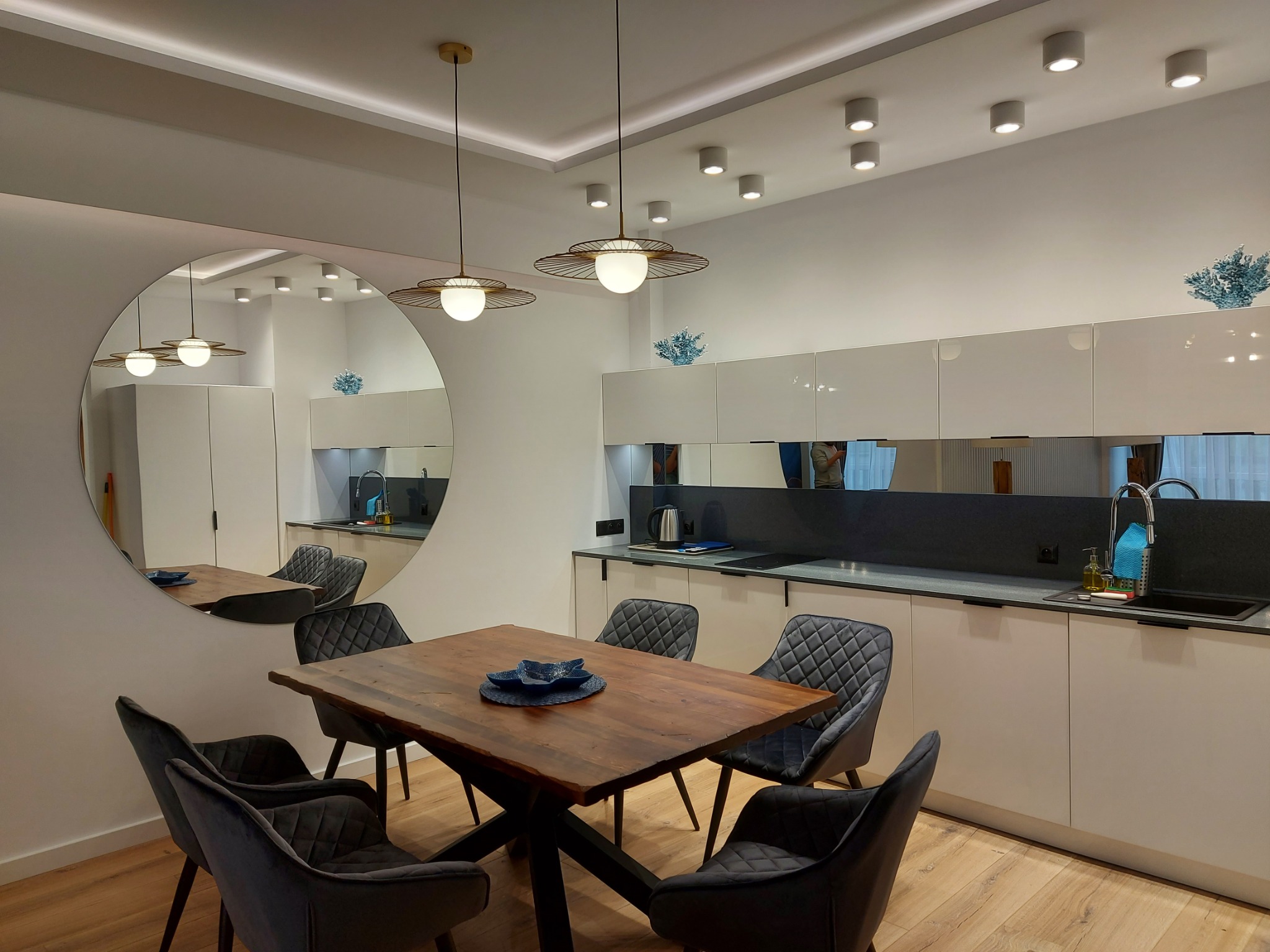 Nowoczesne wnętrze: drewniany stół z dekoracyjnymi naczyniami, szare krzesła z pikowaniem, biała zabudowa kuchenna z czarnym blatem, okrągłe lustro i designerskie lampy wiszące.