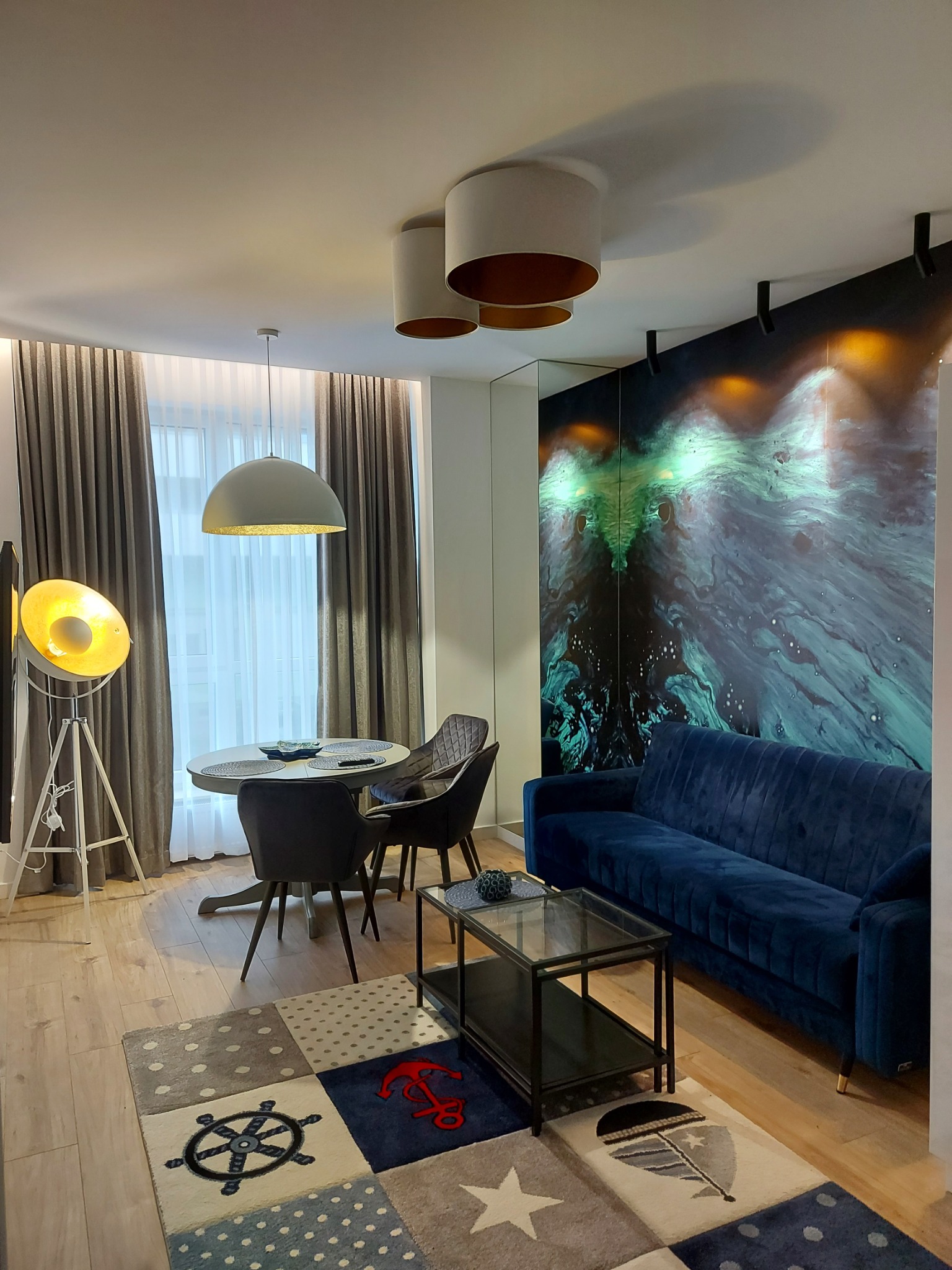 Nowoczesny salon z niebieską sofą, okrągłym stołem i dywanem w marynistyczne wzory, oświetlony lampą studyjną i wiszącą lampą kopułową, z dekoracyjną ścianą z motywem abstrakcyjnych fal.