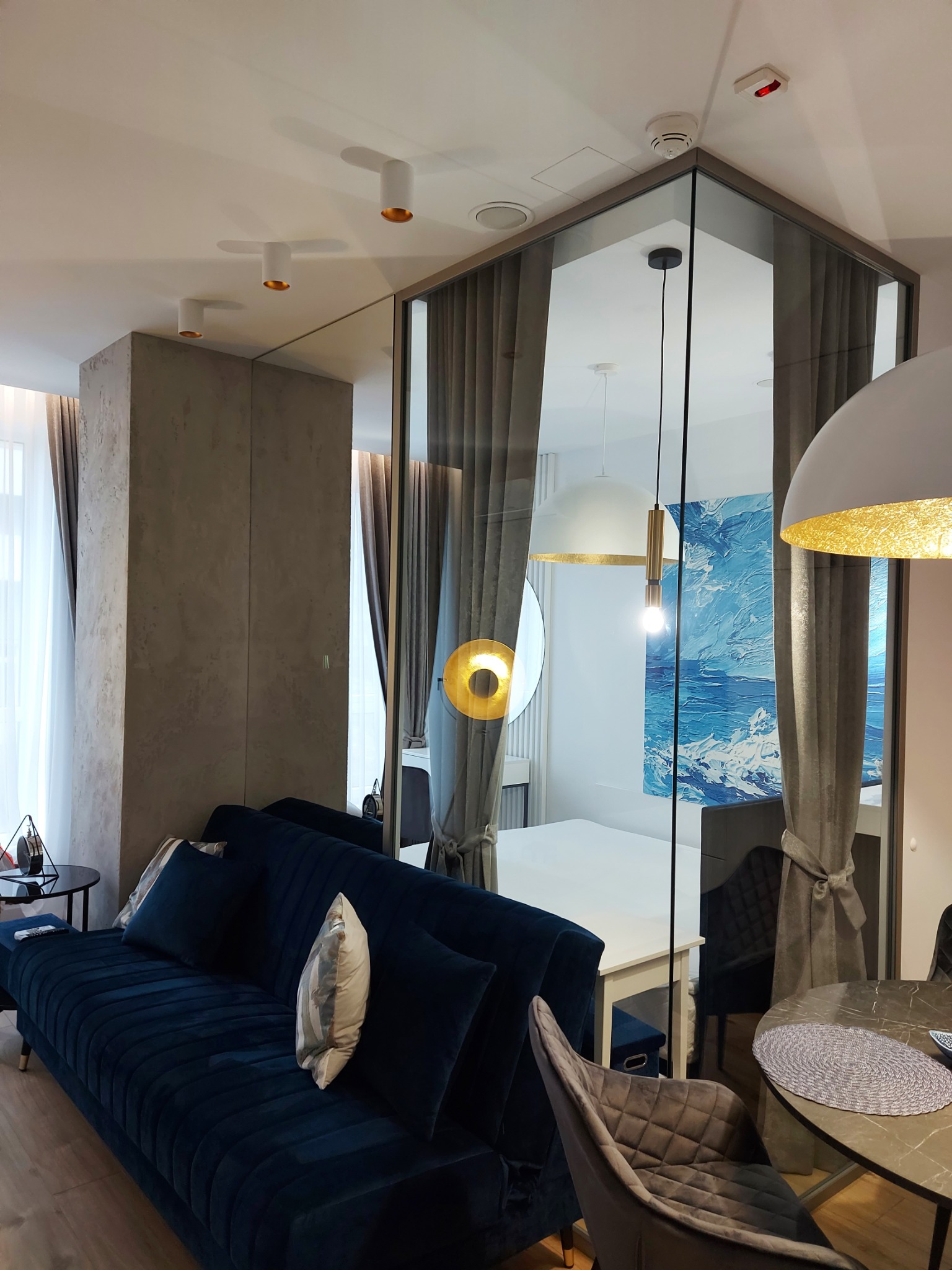 Salon z wydzieloną szklaną ścianą sypialnią, granatowa sofa z poduszkami, szara ściana z efektem betonu, okrągły stół z plecioną podkładką, designerskie lampy sufitowe i wiszące.