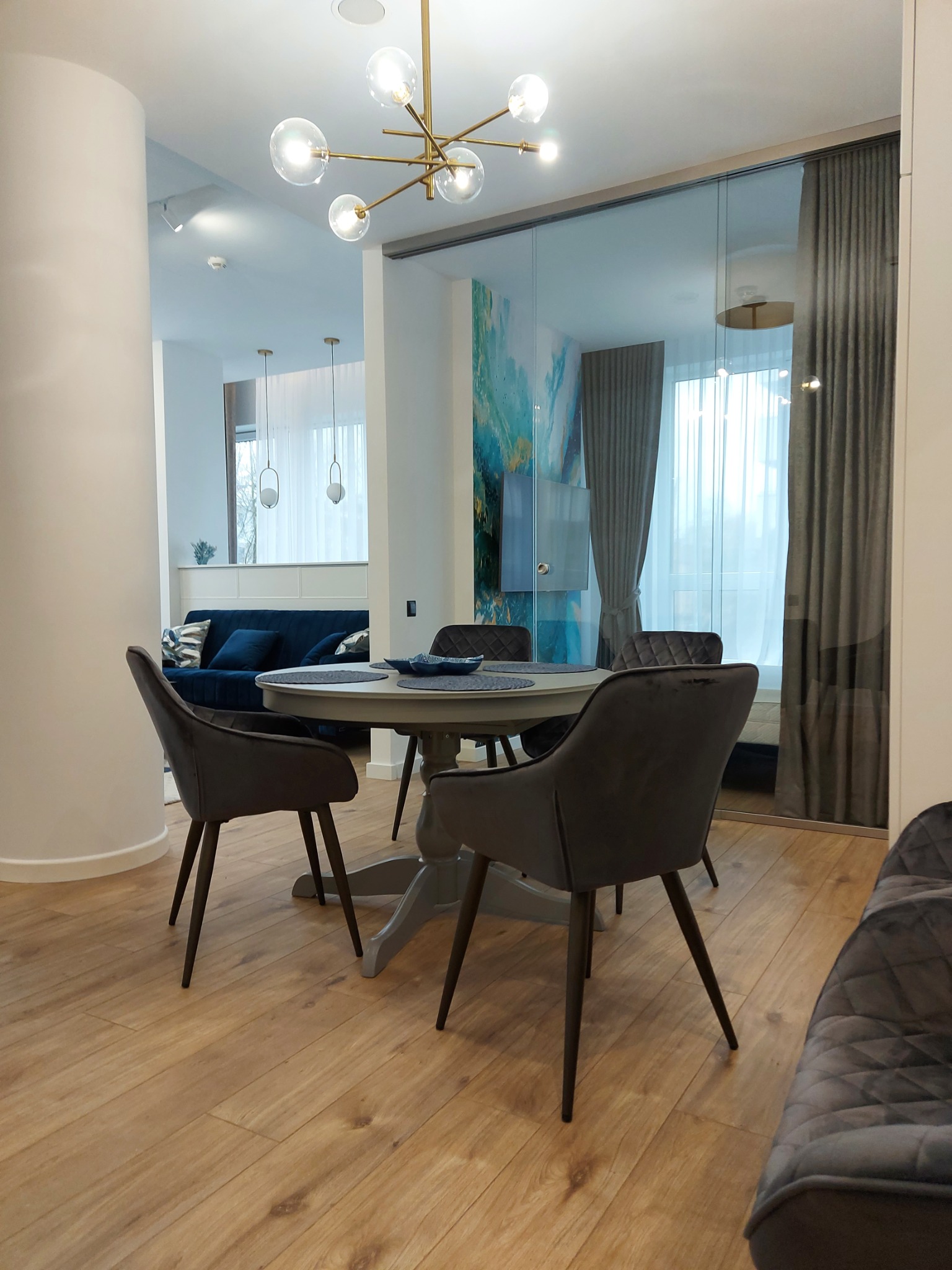 Jasne wnętrze jadalni z okrągłym, szarym stołem i krzesłami na drewnianej podłodze, widok na salon z niebieską sofą i przeszkloną ścianą z zasłonami.