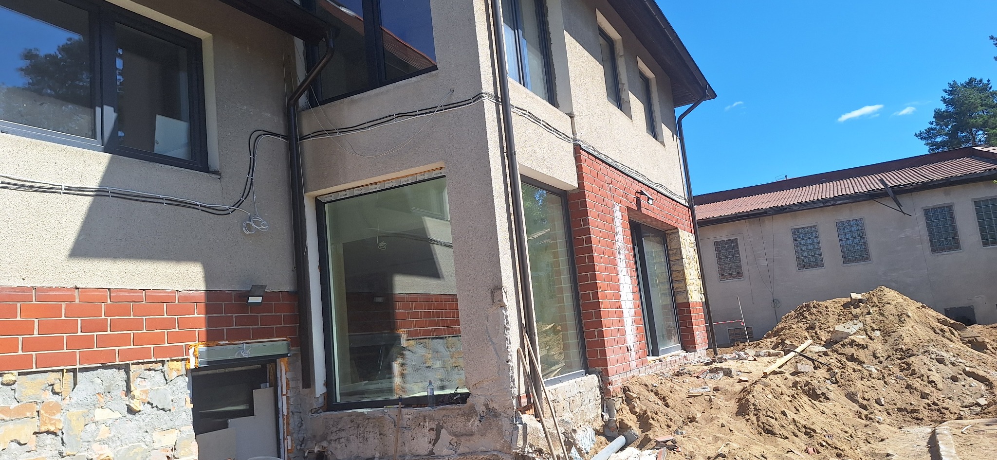Budynek w trakcie prac elewacyjnych z widocznym fragmentem ściany z cegły, nowymi oknami i przygotowanym terenem. Prace w toku, widoczne materiały budowlane.