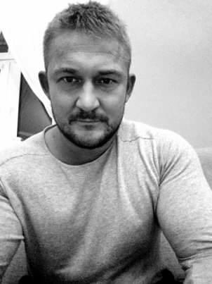 Portret mężczyzny z krótkimi włosami i zarostem, ubranego w szarą bluzę, na rozmytym tle.